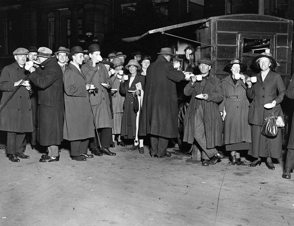 A group of individuals enjoying a leisurely coffee break outdoors. Dressed in elegant long coats and stylish hats, these people exude the timeless charm of a bygone era. This vintage black-and-white photograph evokes nostalgia and a sense of sophistication. Explore the past and savour the moments frozen in time with this remarkable snapshot