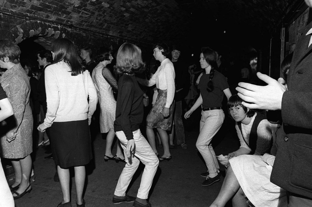 ChatGPT In this monochromatic image, a group of people is captured dancing in a club, with some others choosing to remain seated.