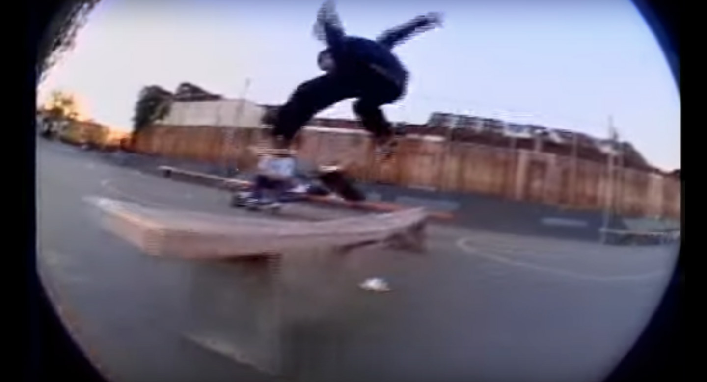 A man flips his skateboard over a bench in a 1990s video.