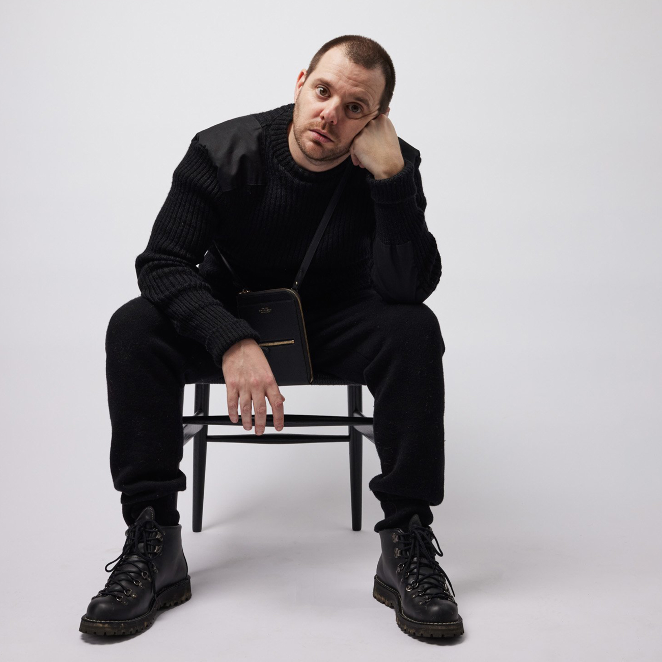 Mike Skinner also known as 'The Streets' sits on a chair wearing all black against a white background in a thinking pose. He is also wearing a small shoulder bag.