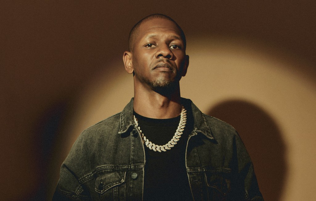 Rapper Giggs stans in front of a beige background with a spotlight shining on him.
