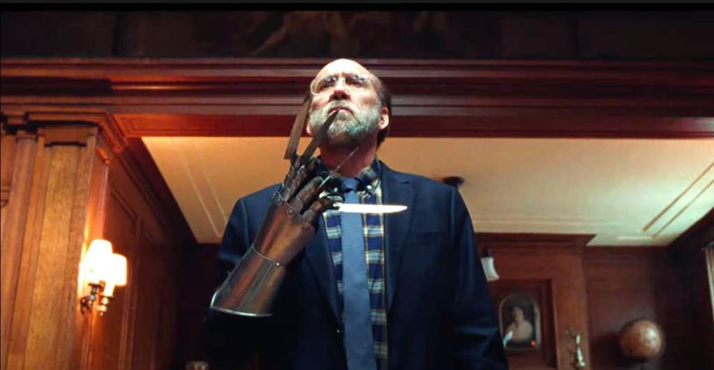 Nicholas Cage stands in a hallway portraying 'Paul' from his new film 'Dream Scenario'. He is wearing an informal blazer with shirt, and is carrying a metal glove with razors on each finger.
