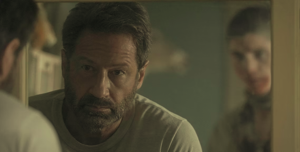 Actor David Duchovny stares into the mirror as a ghostly looking person stands in the background.