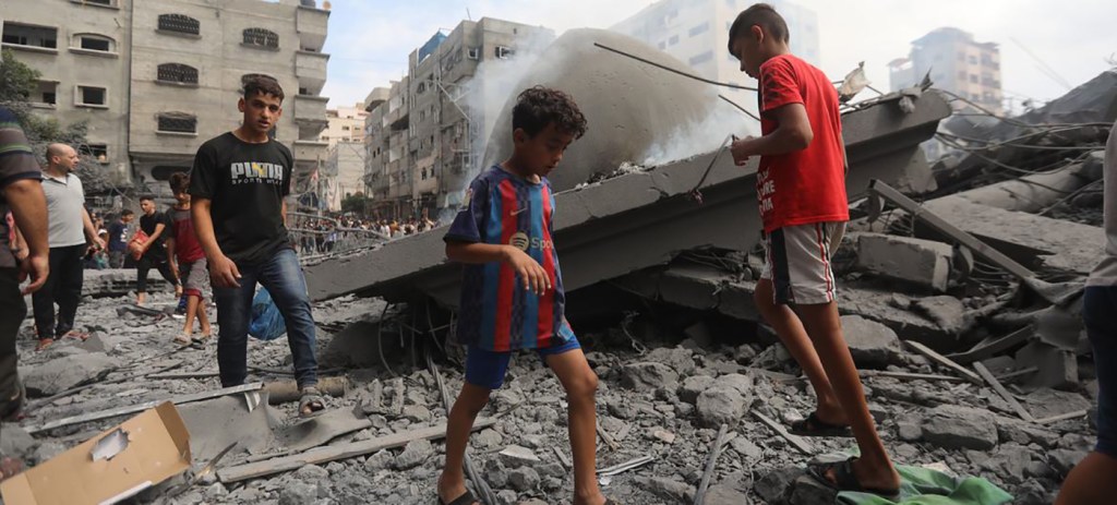 Young Palestinian boys walk amongst the rubble of Gaza where Israeli airstrikes have left 1,800 people dead. They're surrounded by destroyed buildings as smoke billows all around.