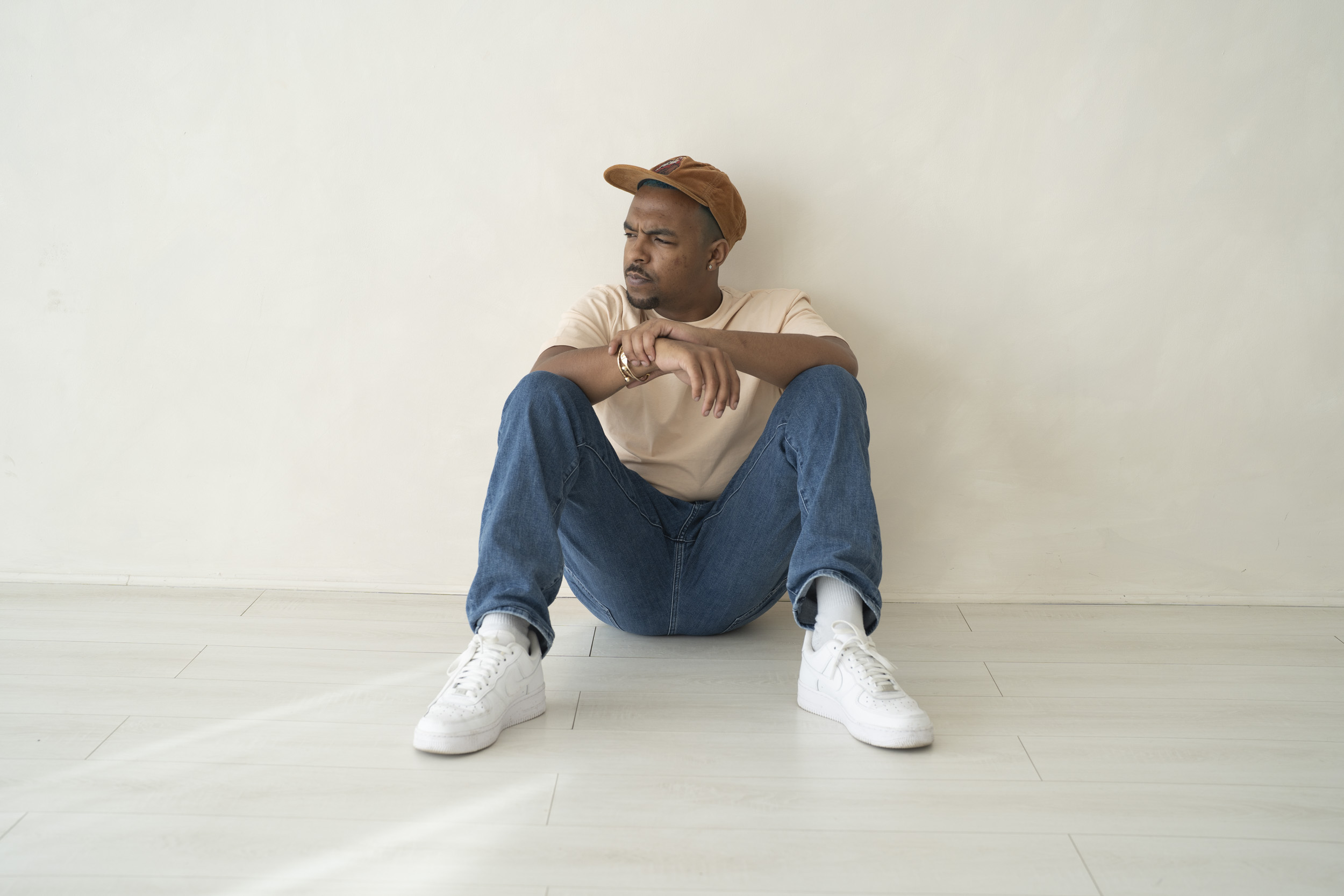 Rapper Karl Sage in a candid shot, wearing blue jeans and white shoes with a brown corduroy cap, against an off-white wall promotional image for his music.