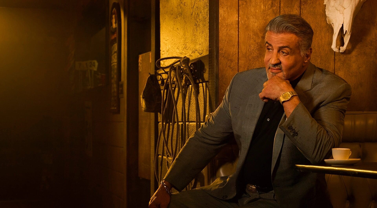 Sylvester Stallone's character, Dwight "The General" Manfredi, sits on a chair, smiling under a dim overhead light.