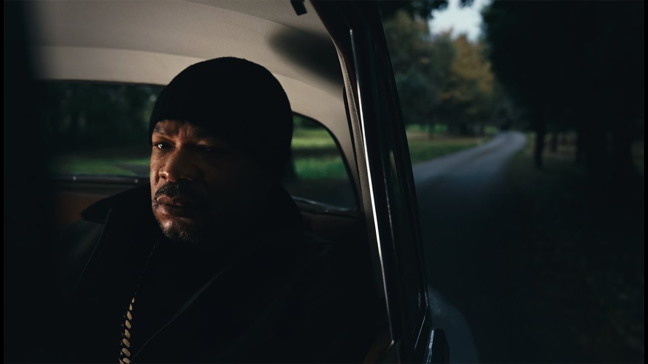 Rapper Xzibit sits in the back of a car, contemplating life. In the background, trees begin to blur as the car moves forward away from them. A window is rolled down slightly in a dark and moody composition, lit only from the inside of the car.