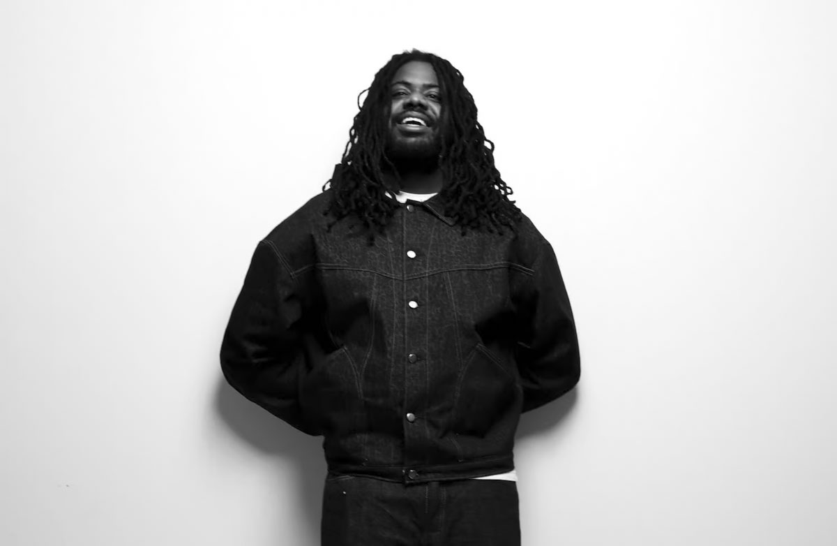 Rapper MIKE smiling in front of a white wall, wearing a denim jacket. Hip-hop artist portrait with a casual and confident look.