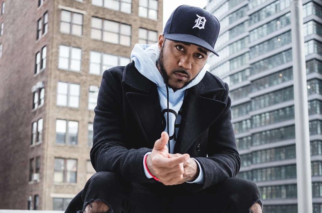 Jason Martin sits down in front of two buildings, wearing a black coat, hooded jumper, and baseball cap.