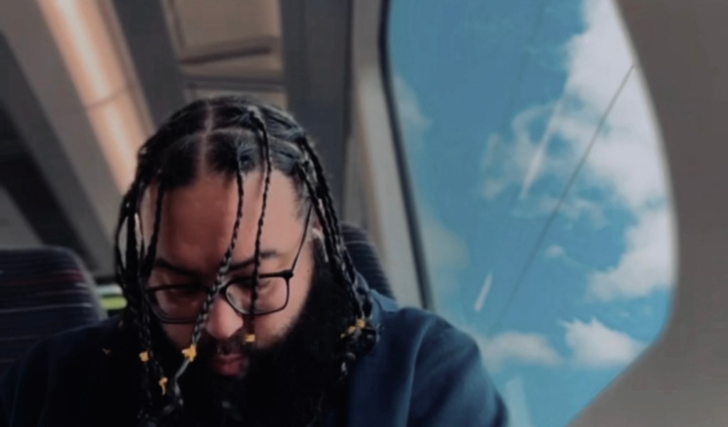 UK rapper Scrulove sitting by a window on a train, contemplating for press still of new single 'Glory' from his 2025 album.
