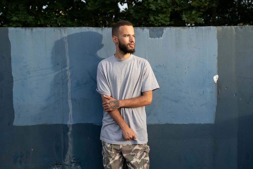 UK hip-hop artist, Sincerely Keane, stands against a blue backdrop wearing a grey t-shirt on a sunny day at golden hour.