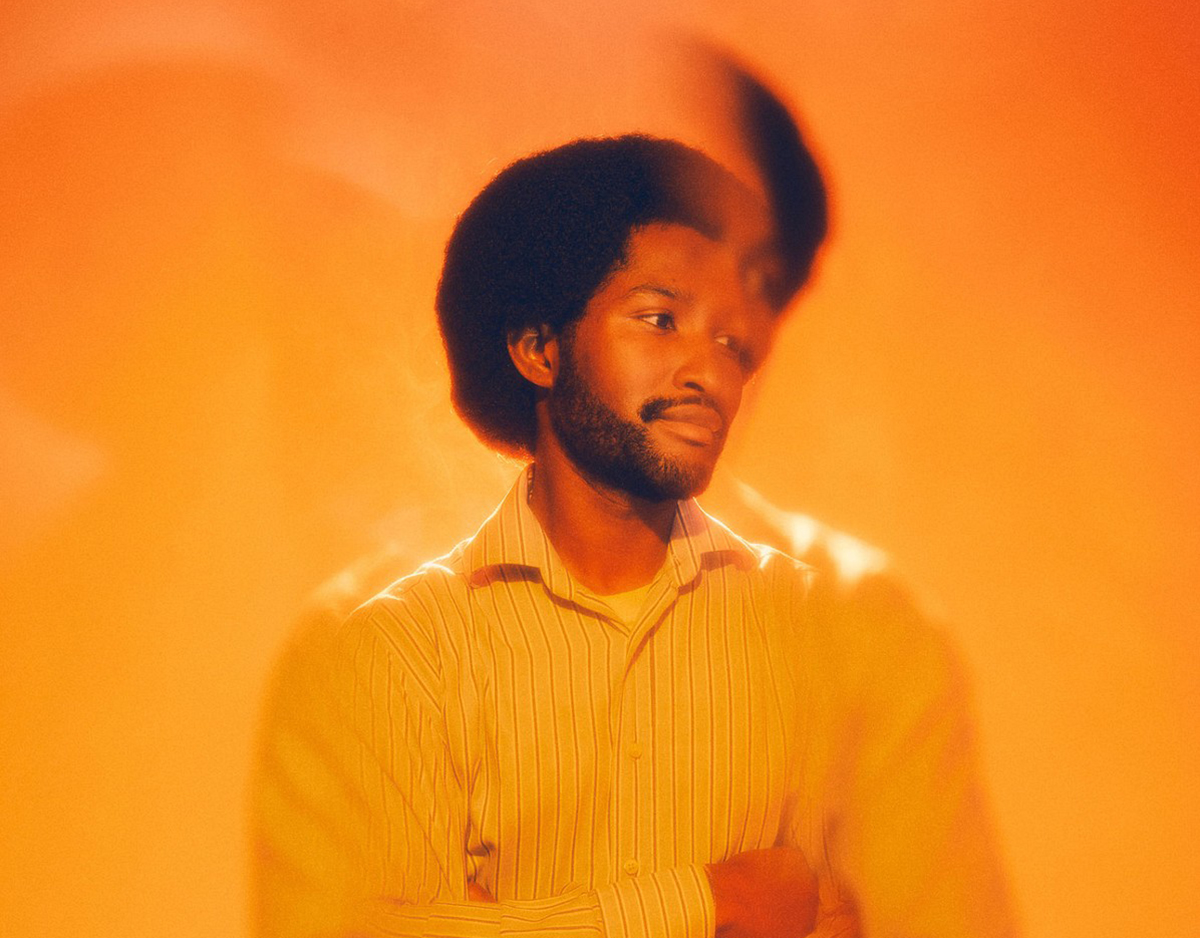Wilfred Cisse in white shirt and brown trousers against orange background, looking left.