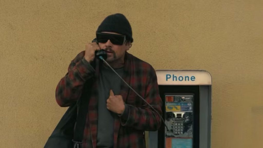 Actor Leonardo DiCaprio sports a long moustache while talking into a pay phone wearing large sunglasses and a red patchwork dressing gown. He is also wearing a black woolly hat.