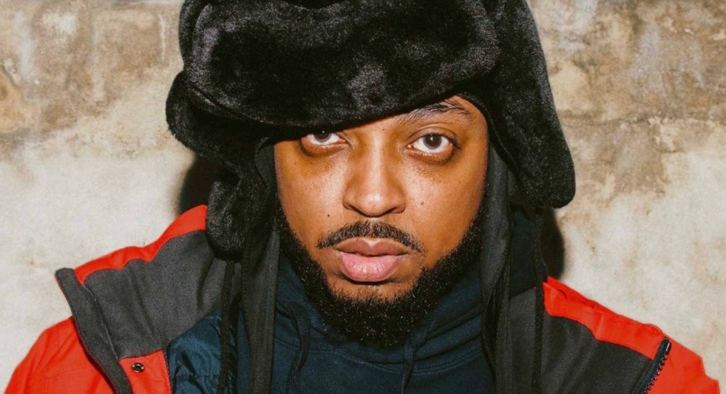 Rapper Da Flyy Hooligan stands against a brick wall, wall wearing a black hat and red jacket.