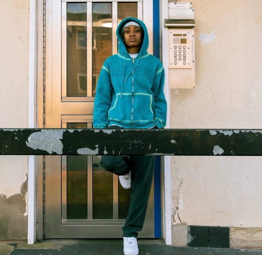Rapper LIV wearing a blue hooded jumper and green trousers posing in an urban street scene