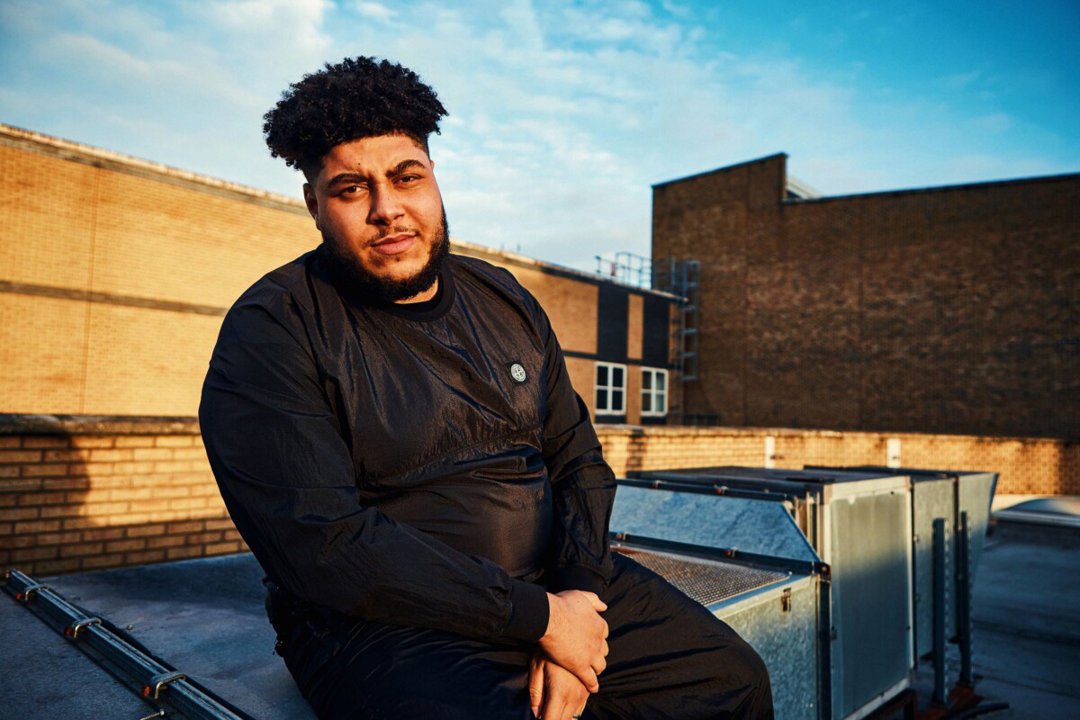 Big Zuu sits on a rooftop at sunset in London. He is wearing a black sweater and trousers.