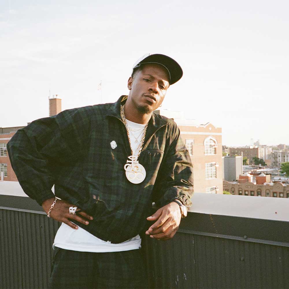 Rapper Joey Badass stands on top of a rooftop at sunset in New York City wearing a baseball cap and jacket.