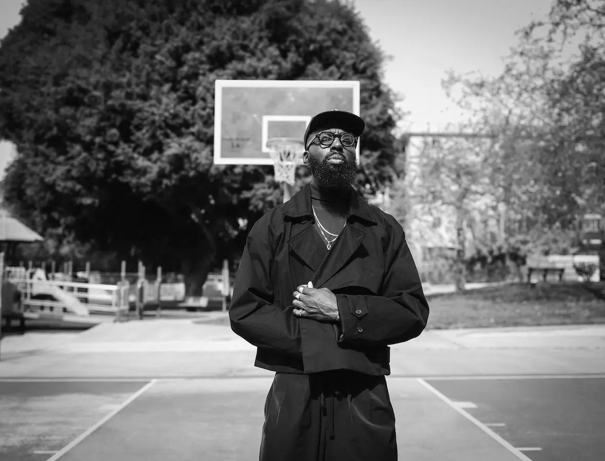 Made Allayne stands in a basketball court wearing a black outfit, baseball cap, and spectacles.