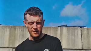 Welsh rapper Chrxn wears a black t-shirt against a garden fence and blue skies.