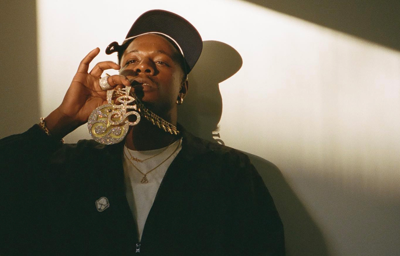 Joey Badass wears a baseball cap and holds a medallion as soft light enters the room filled with shadows.