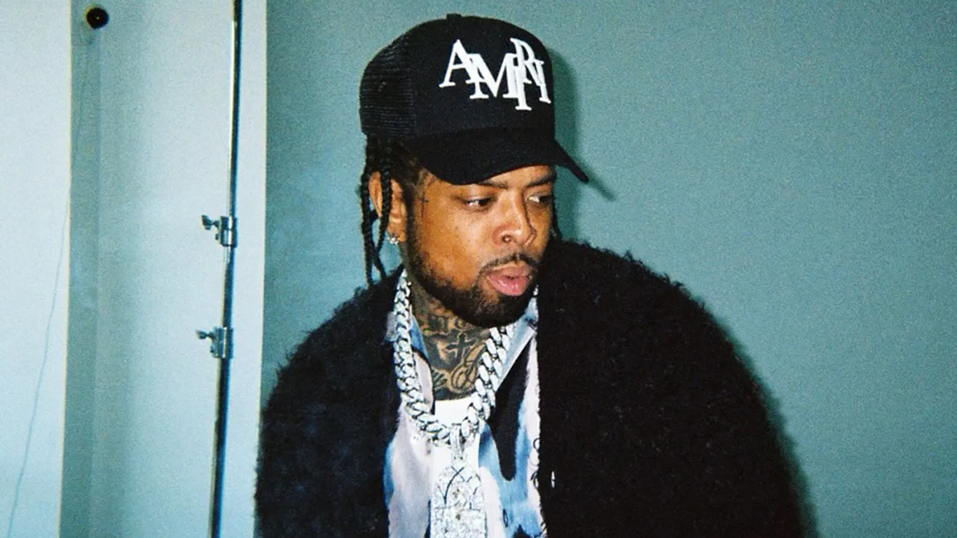 Westside Gunn wearing a black jacket, baseball cap, and platinum diamond chain, standing against a turquoise wall.