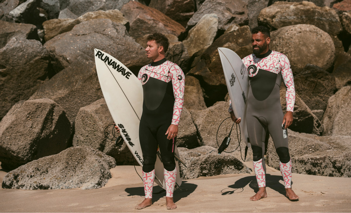 Two surfers stand on a beach for the Pink Onda Project.