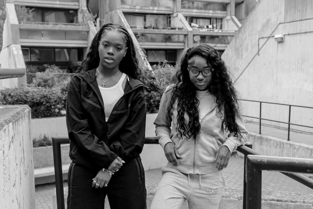 Rappers Cristale and TeeZandos in monochromatic outfits against a brutalist backdrop.