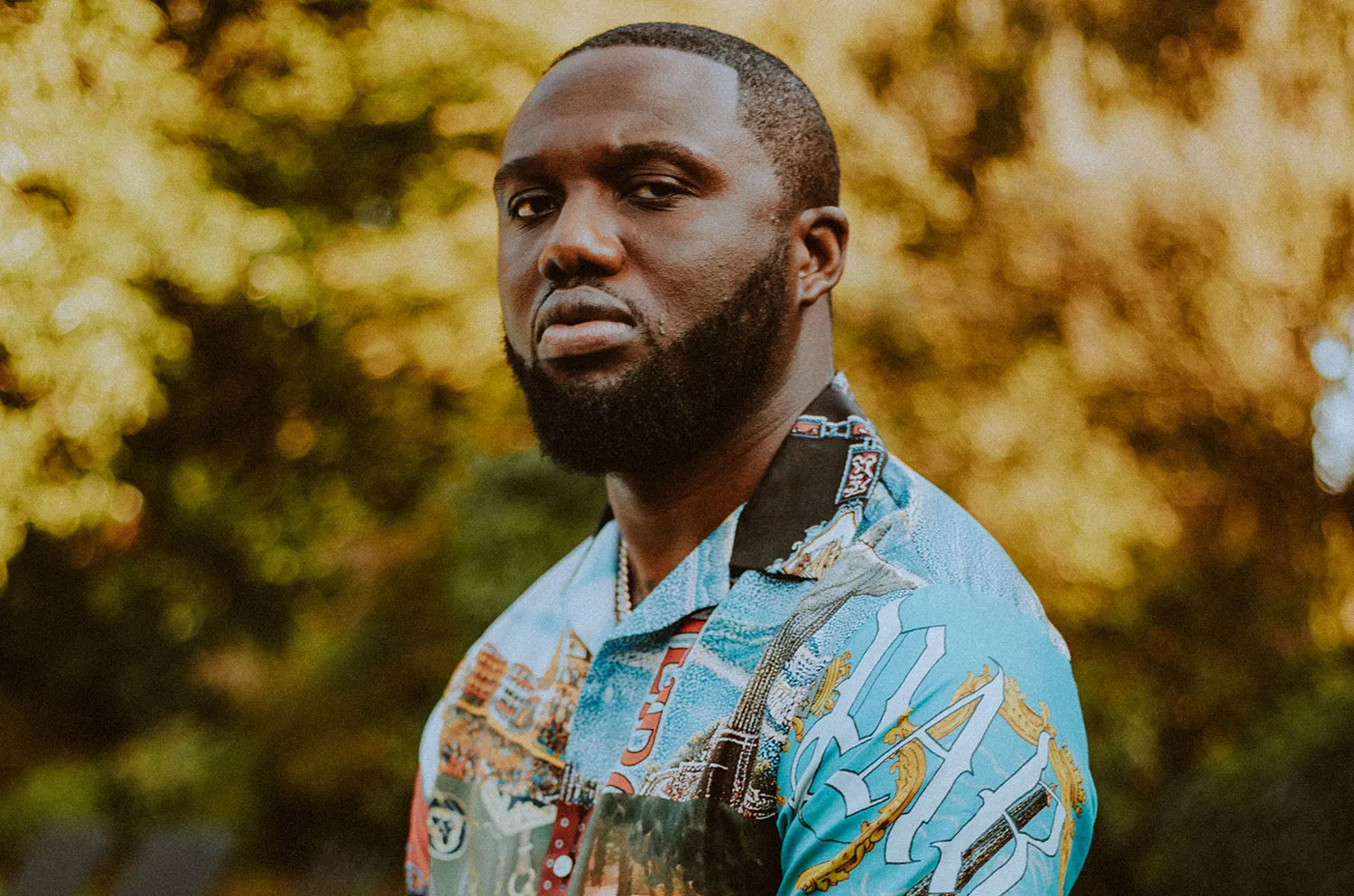 Headie One stands in front of trees, wearing a blue shirt at sunset.