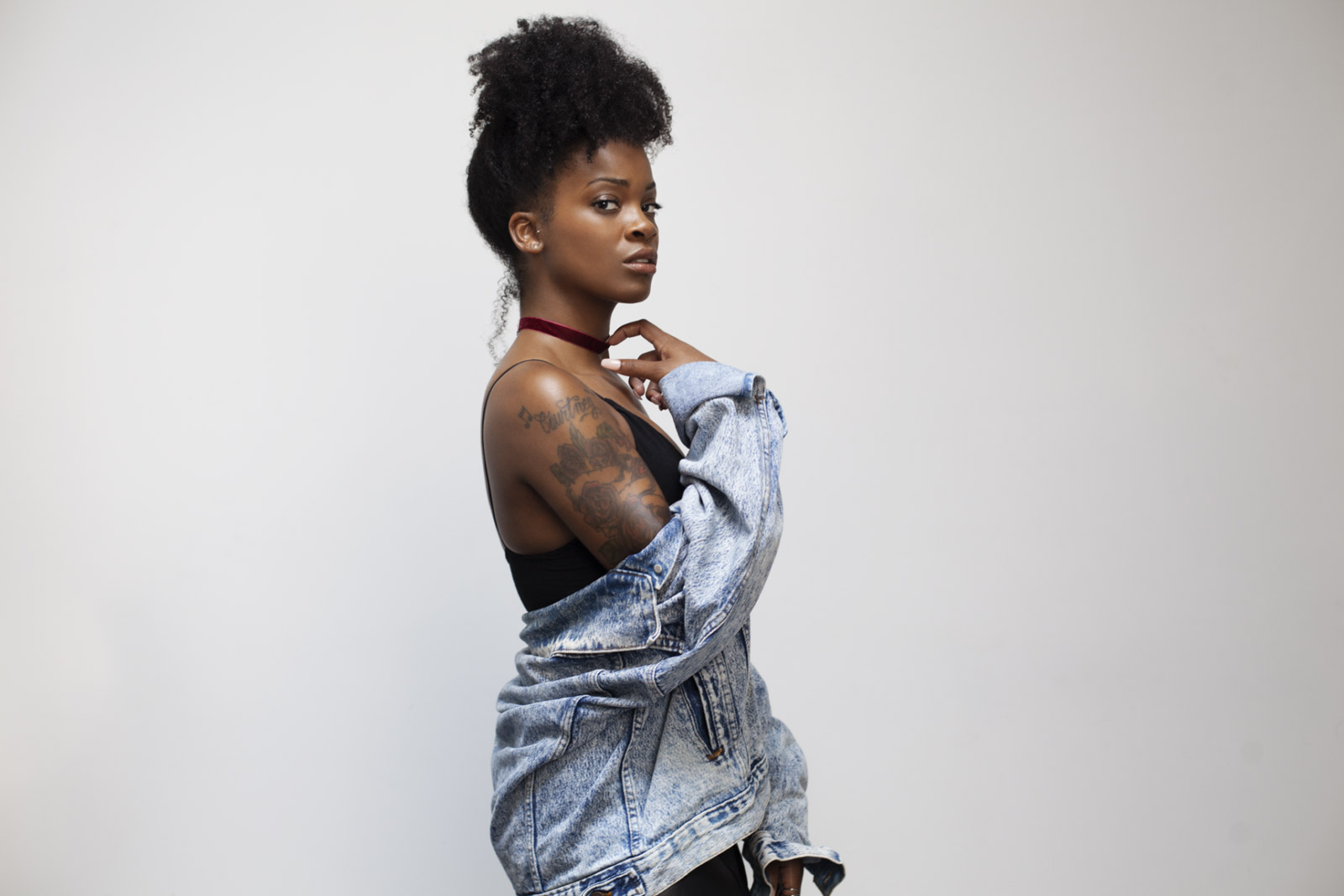 Ari Lennox wearing a denim jacket draped off her shoulders, looking confidently at the camera.