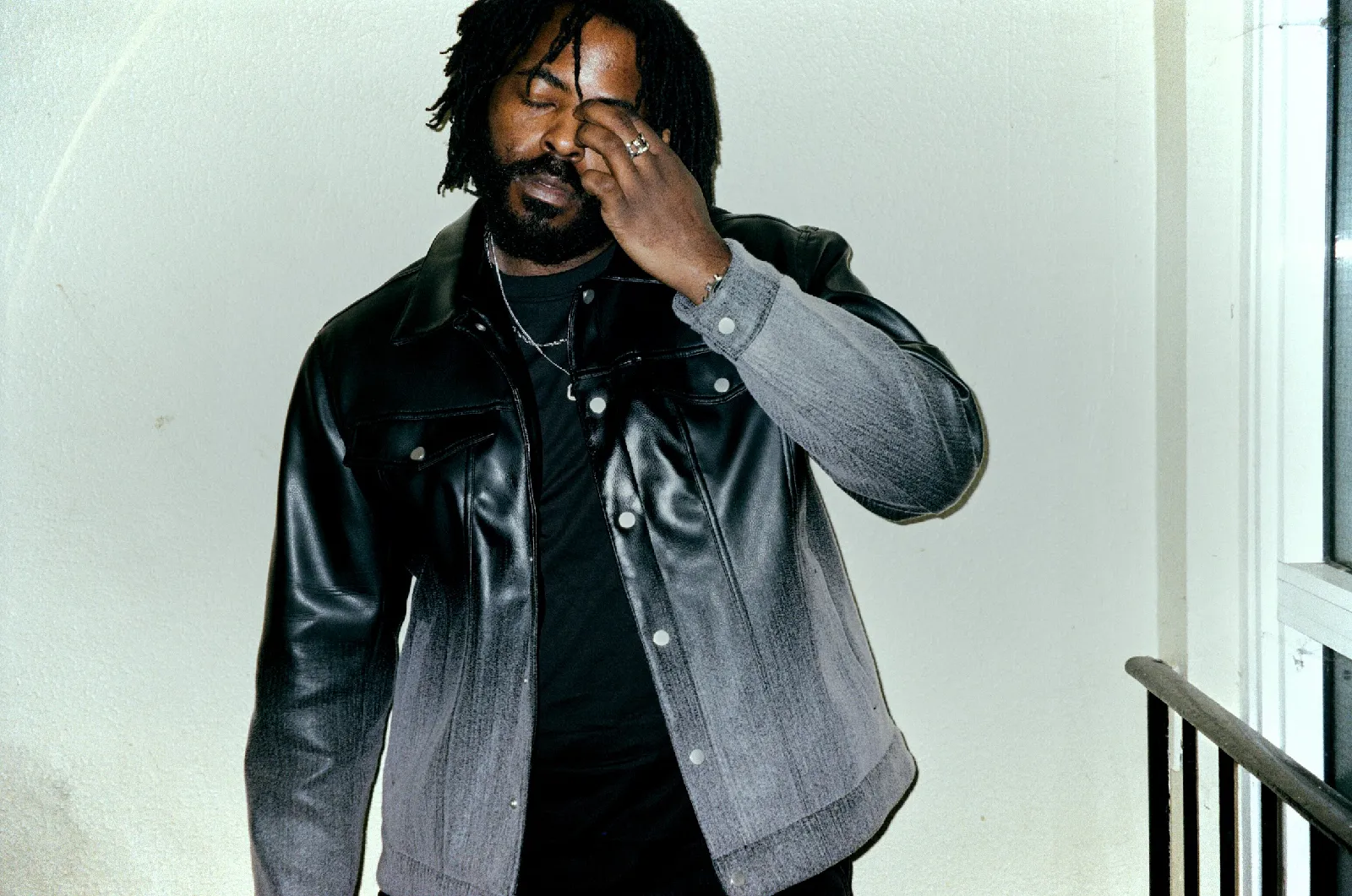 UK rap artist iiiBerealz stands in a hallway wearing a denim shirt, rubbing his eye.