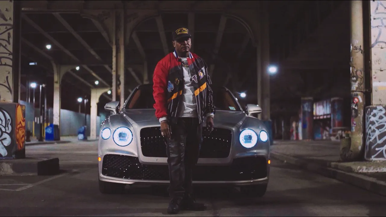 AZ stands in front of his luxury car, wearing a baseball jacket, in a promotional still for his new music video for “Uniqueness."
