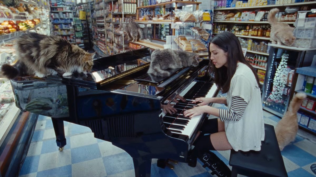 Olivia Rodrigo playing a piano inside a grocery store with two cats in a scene from her music video.
