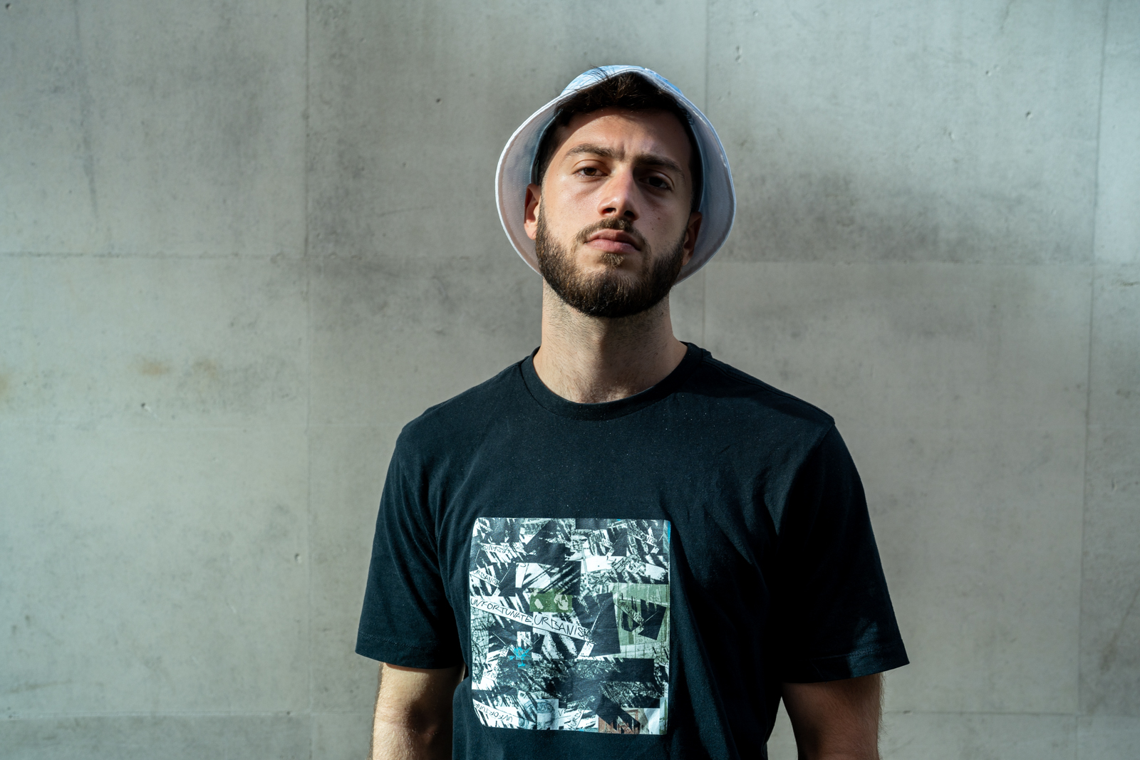 UK rapper The Philosopher standing against a gray concrete wall, wearing a white bucket hat and black t-shirt.