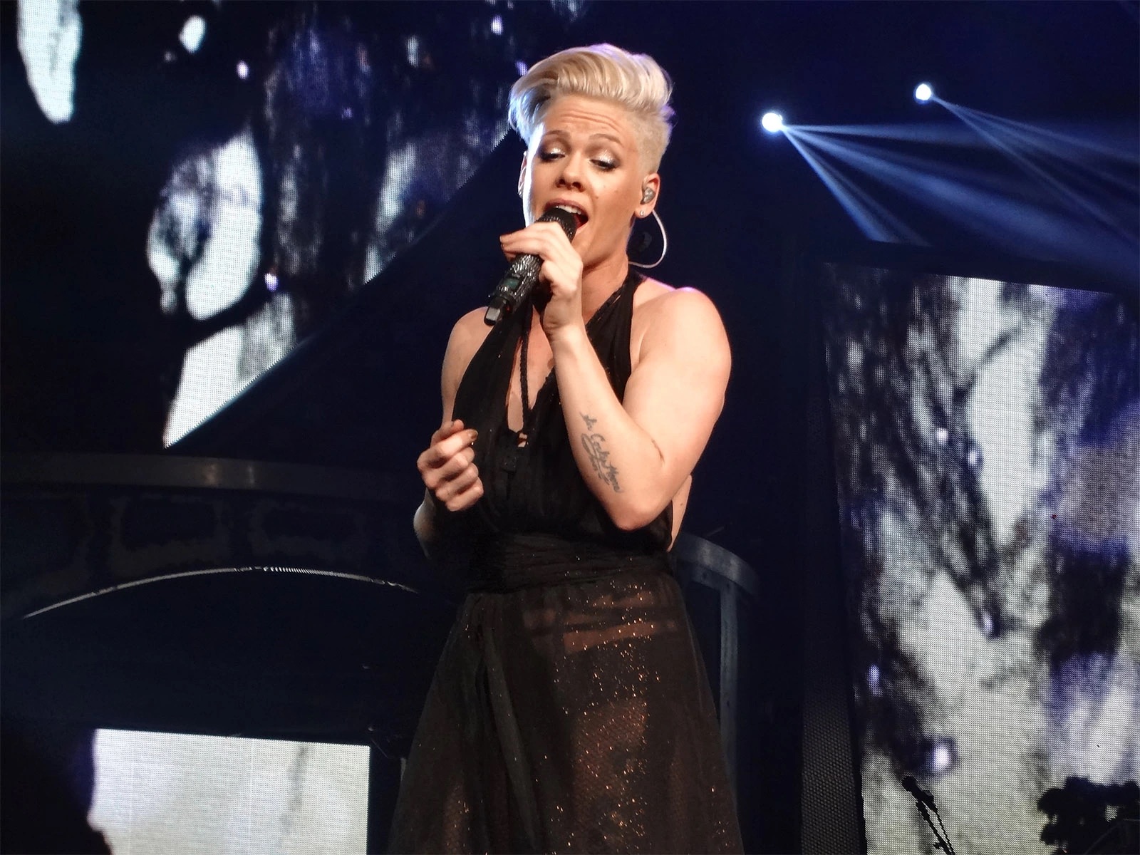 Pink performing on stage in a black dress, singing into a microphone under concert lights.