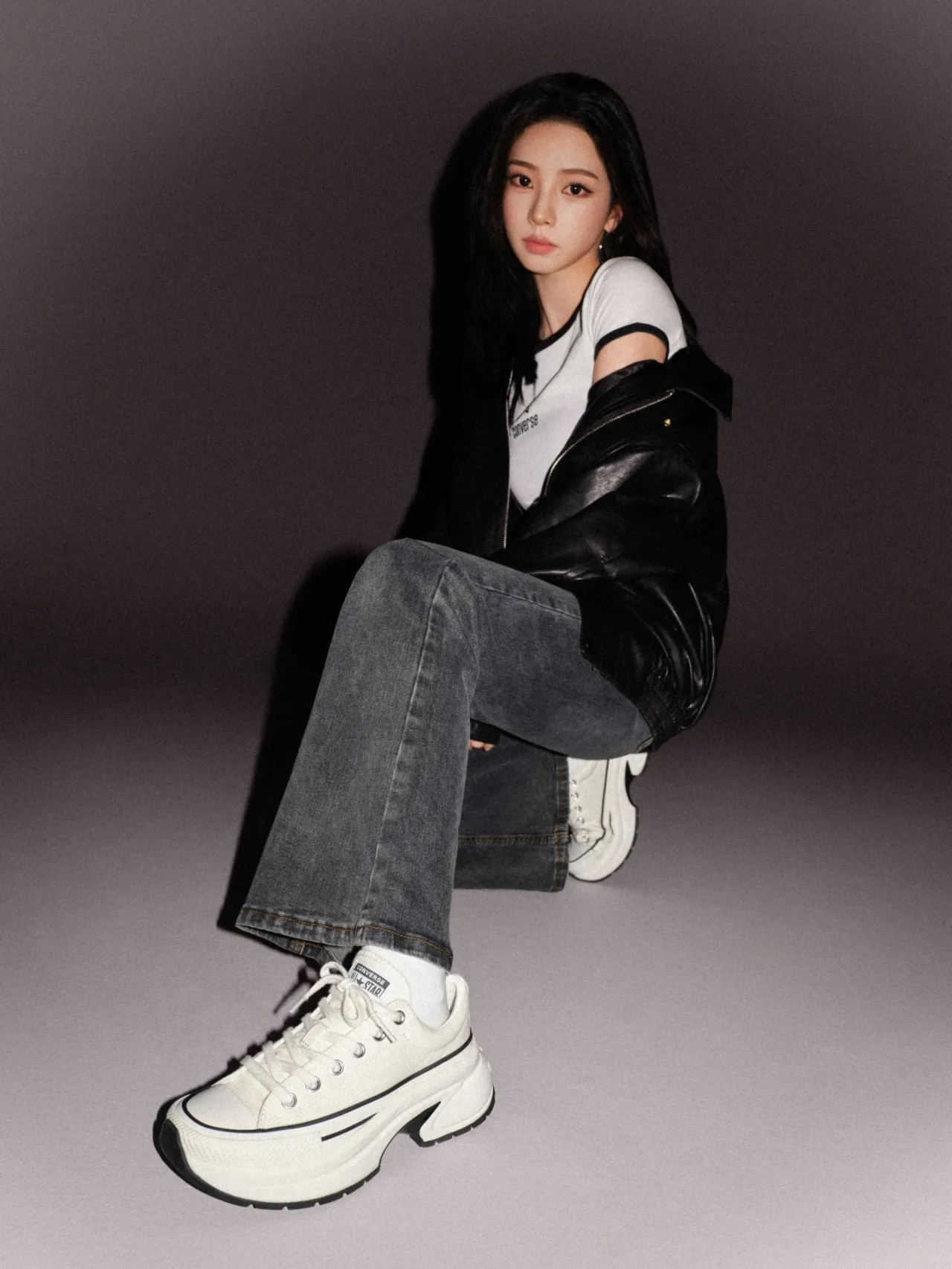Karina poses seated in a Converse campaign wearing a white Converse T-shirt, oversized black leather jacket, grey jeans and chunky white Converse sneakers.