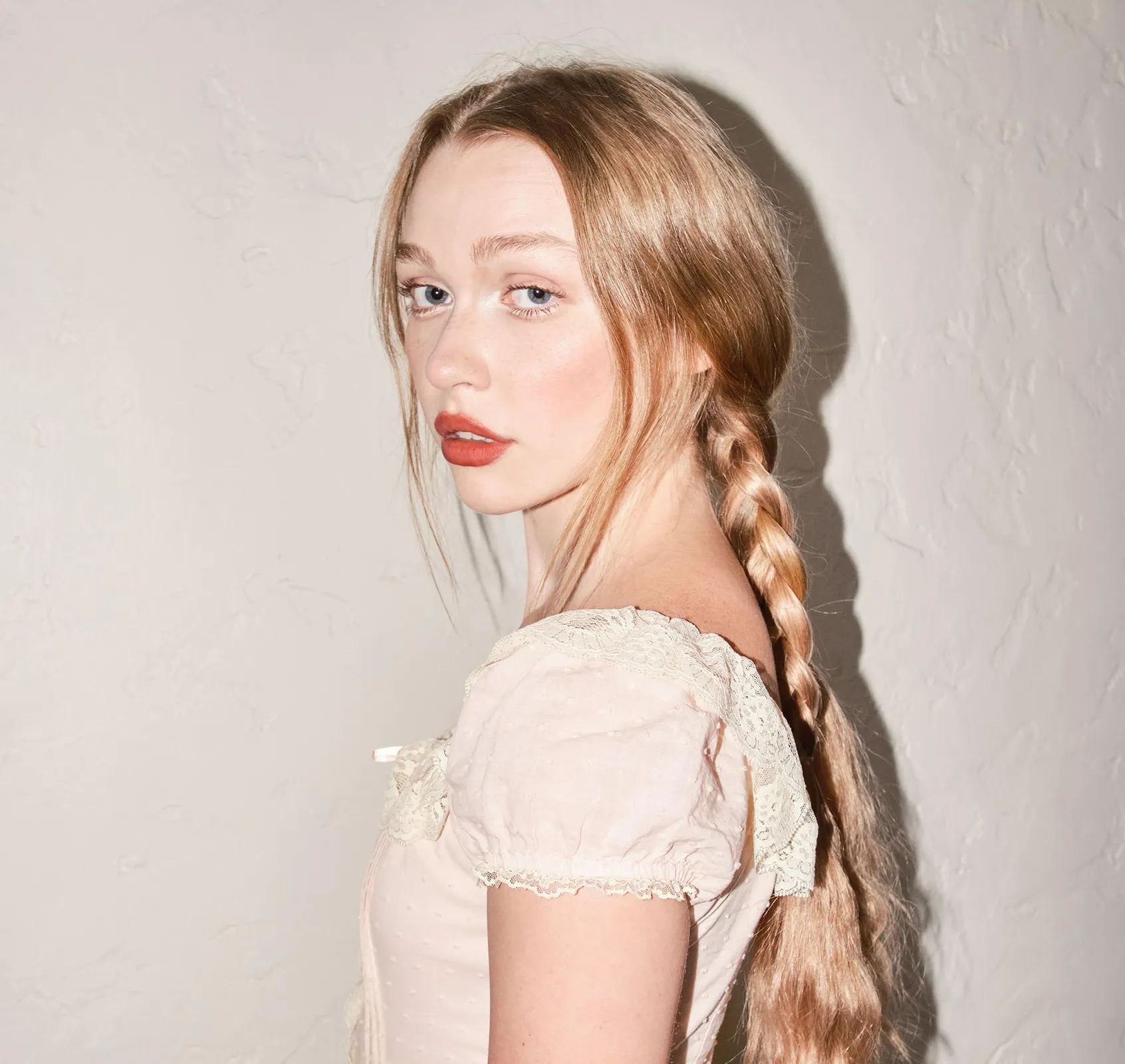 Mollie Elizabeth pictured in a pale pink vintage-style dress, looking over her shoulder against a textured wall.