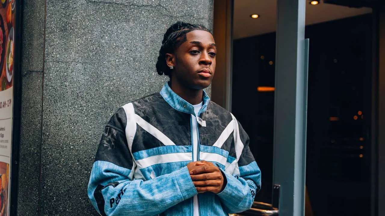 WhyDee wearing a blue jacket with his hands clasped against a grey concrete wall.