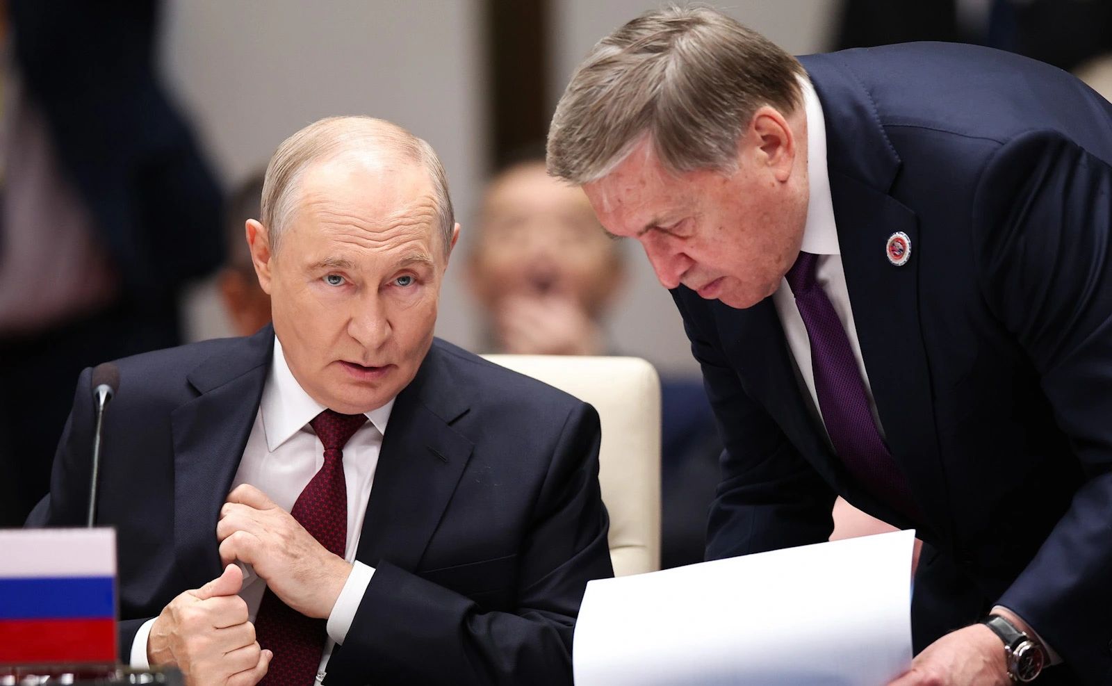 Vladimir Putin seated at a Shanghai Cooperation Organisation summit as aide Yuri Ushakov leans towards him holding documents.