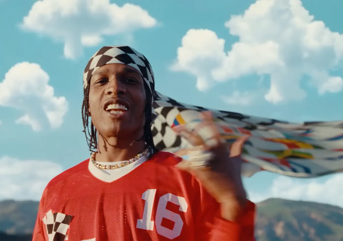 A$AP Rocky wearing a red football jersey and headscarf against a pale blue sky and mountains from the music vdieo of "SHROOM CLOUDS."