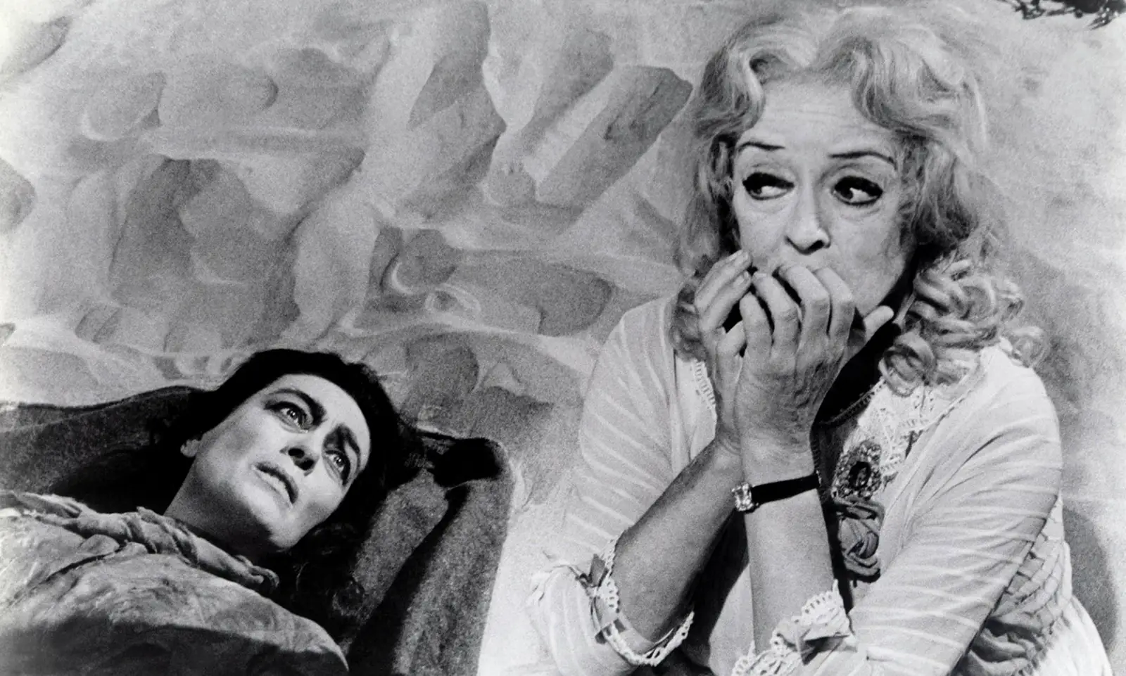Black-and-white promotional still from What Ever Happened to Baby Jane? (1962), featuring Joan Crawford lying in bed while Bette Davis sits beside her, anxiously covering her mouth with her hands.