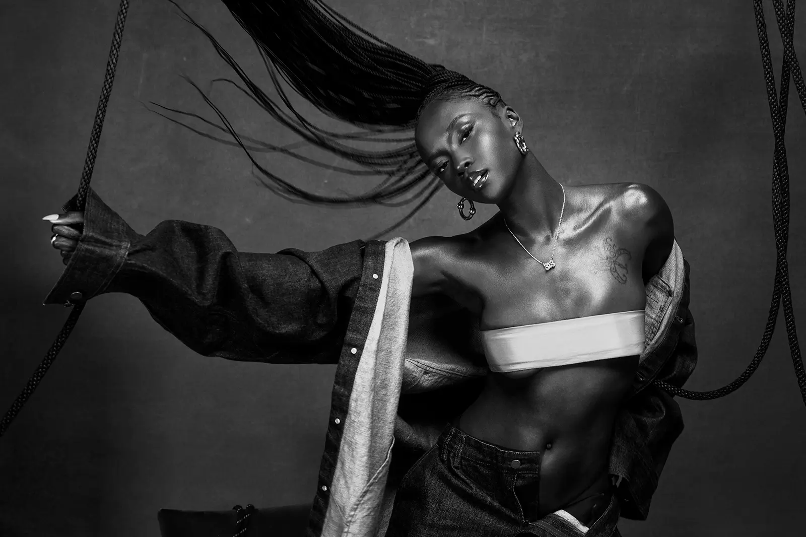 Black-and-white portrait of R&B singer Ebony Riley with long braids sweeping overhead, wearing a bandeau top and an open denim jacket while holding a rope.