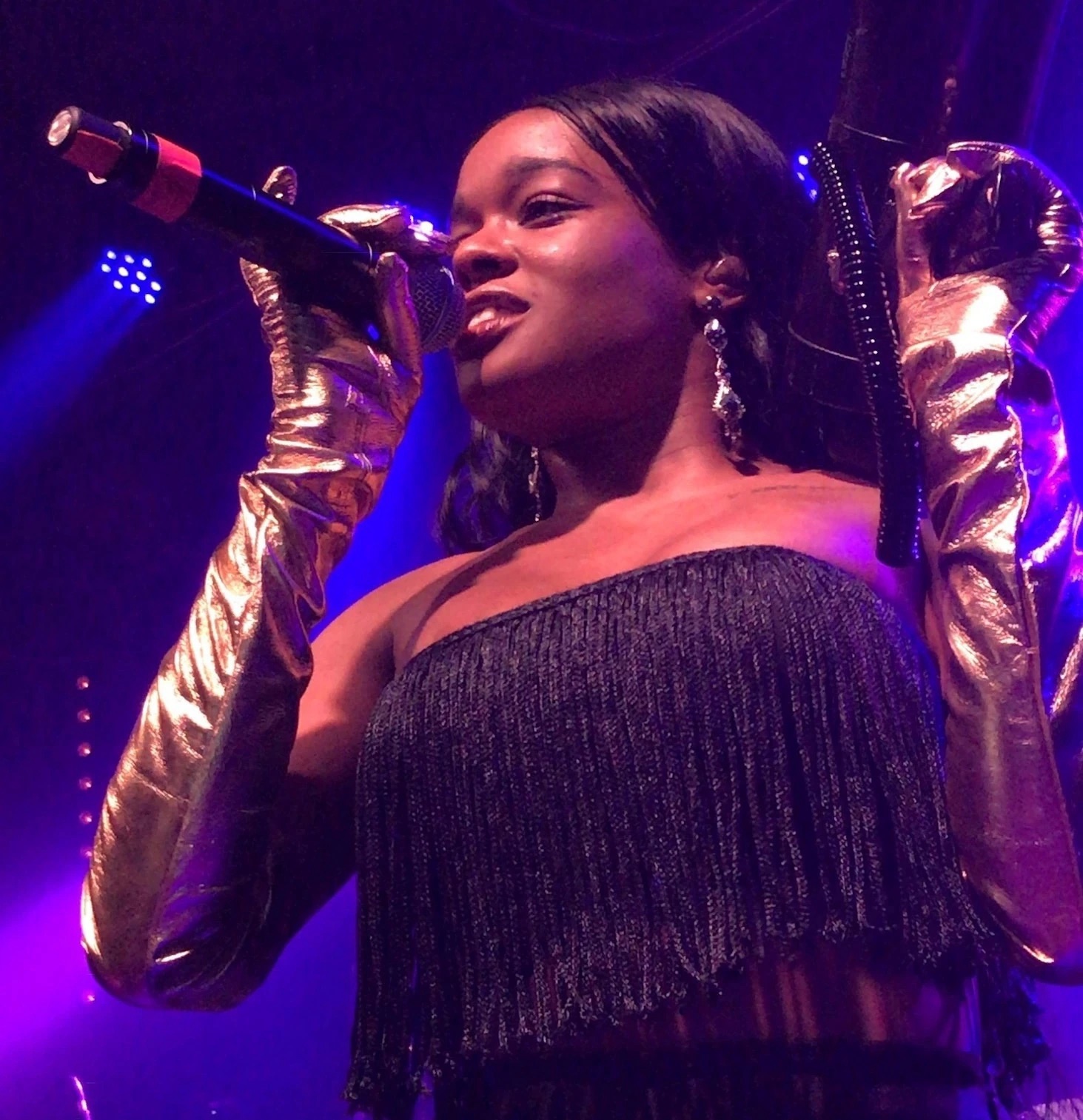 Azealia Banks performing on stage in a black fringed outfit and gold gloves under purple lighting.