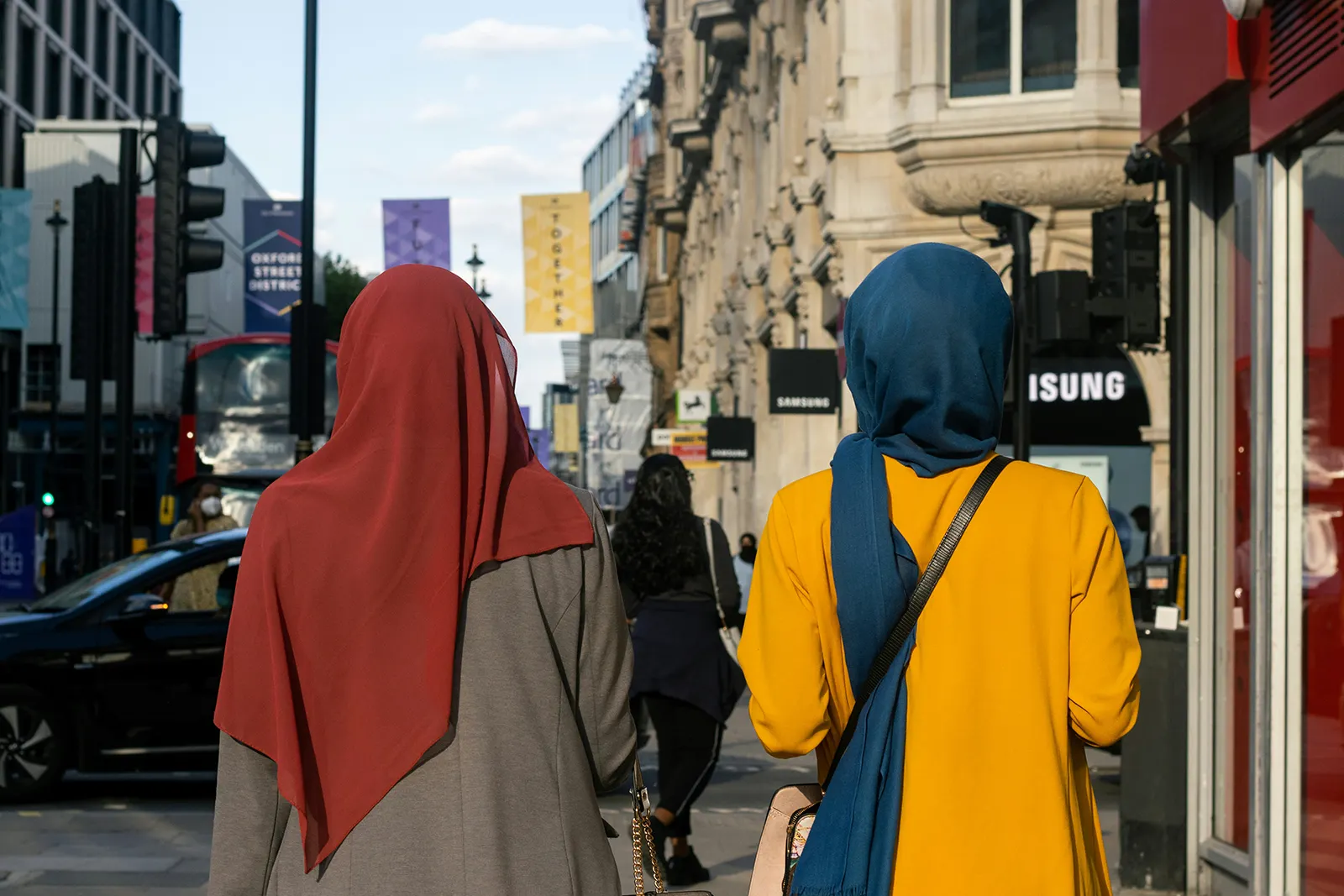 Two Muslim women wearing hijabs walk along Oxford Street in London, with shops, traffic and pedestrians in the background.