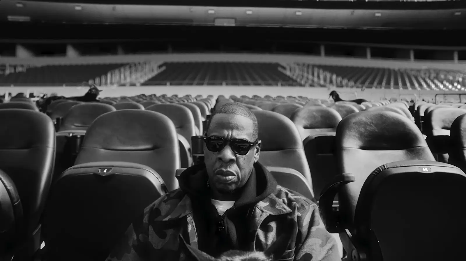 Jay-Z wearing black sunglasses and a camouflage jacket in a promotional image for the 2027 Super Bowl halftime show, seated in an empty stadium.