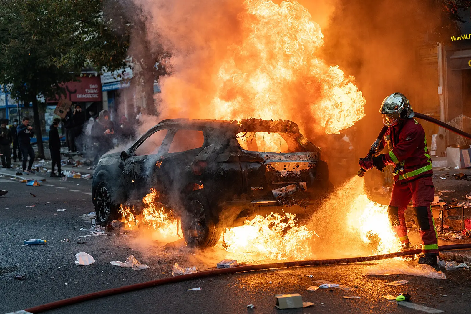 A firefighter wearing red protective gear attempts to extinguish a burning car as towering flames and thick smoke fill a Paris street, with bystanders visible in the background.