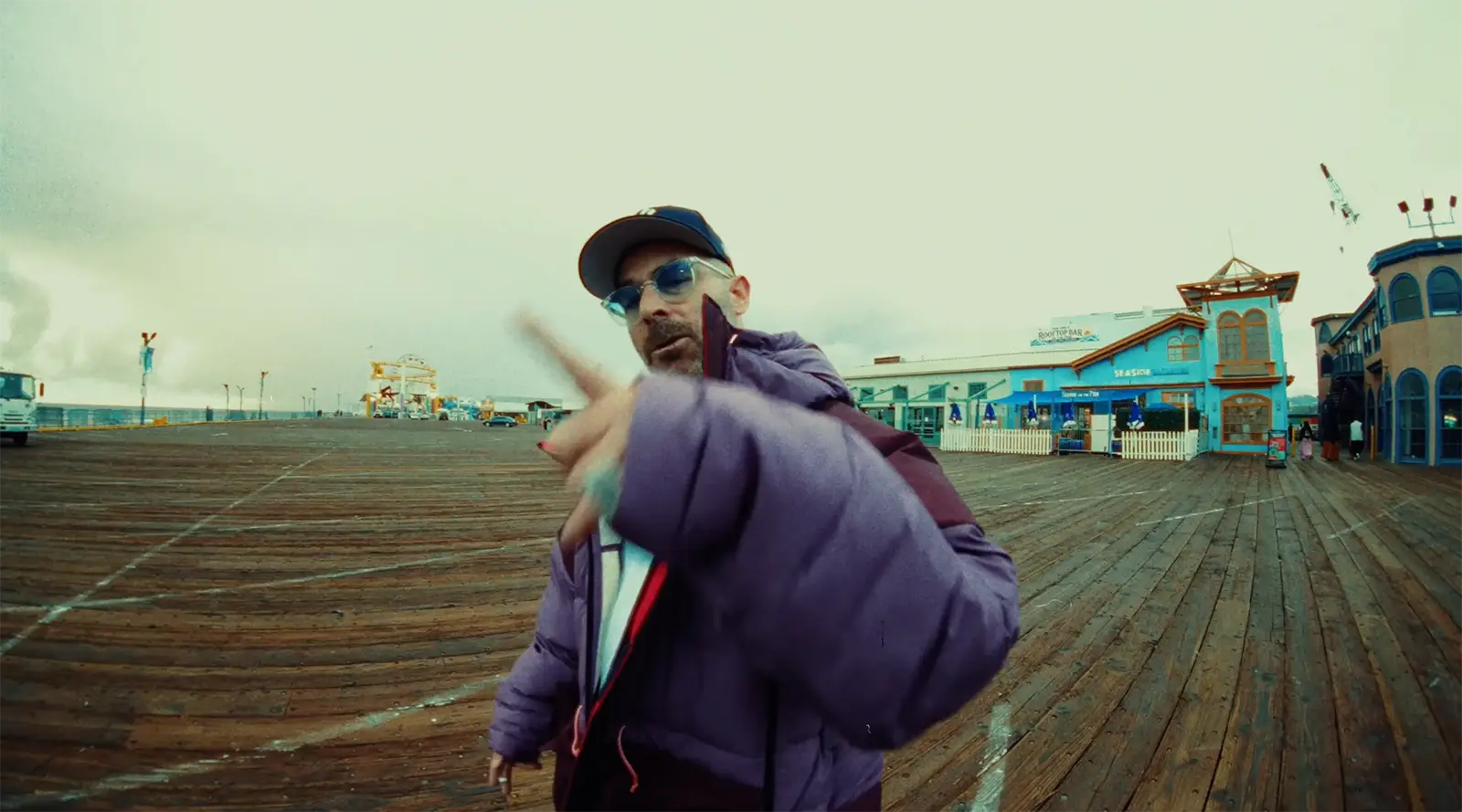 The Alchemist raps wearing a purple puffer jacket, baseball cap, and sunglasses on a pier.